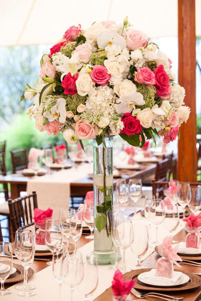 Tall glass vase filled with roses, hydrangeas, and orchids as a floral centerpiece on a beautifully set wedding table with glassware and pink accents.